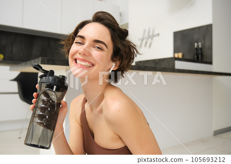Smiling active young woman in sportswear, sits on yoga mat with water bottle, drinks stays hydrated during workout training session from home, fitness in the house on floor Smiling active young woman in sportswear, sits on yoga mat with water bottle, drinks stays hydrated during workout training session from home, fitness in the house on floor 105693212