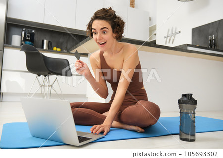 Image of young fitness girl, female athlete at home, workout on kitchen floor, holding credit card with laptop, purchasing online training session, gym application for home workout 105693302