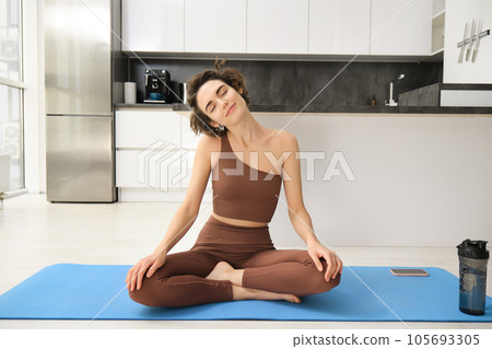 Photo of focused young woman in sportswear stretching her neck while working out on mat at home, doing fitness exercises 105693305