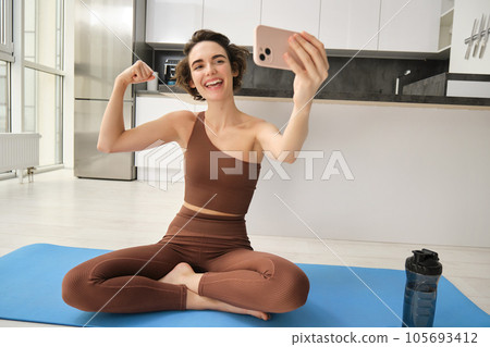 Image of young fitness girl blogger, records her workout at home, doing workout and showing muscles to telephone camera. Woman with smartphone, joining remote yoga training via mobile phone 105693412