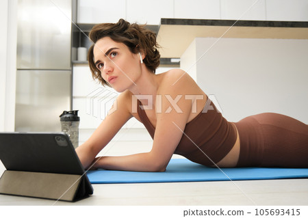 Portrait of fitness girl with thoughtful face, lying on rubber yoga mat at home, listening and watching sport workout video tutorial on tablet, thinking 105693415