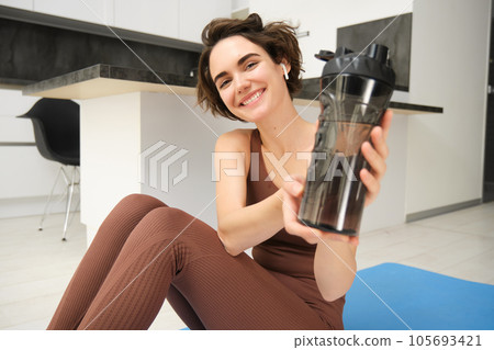 Smiling sportsgirl, woman after fitness training, gives you water bottle, stays hydrated after workout, sits on fitness yoga mat at home on floor 105693421