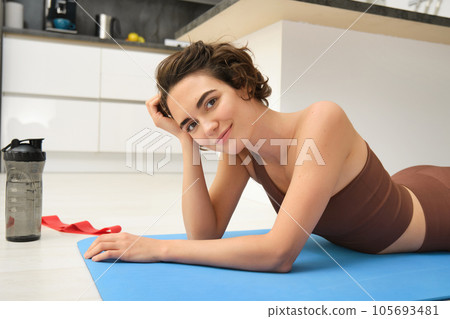 Portrait of fitness girl lying on rubber mat, warm-up before workout, doing pilates exercises at home, drinking water and doing sports, smiling at camera Portrait of fitness girl lying on rubber mat, warm-up before workout, doing pilates exercises at home, drinking water and doing sports, smiling at camera 105693481