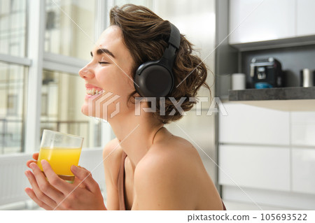 Sport and healthy lifestyle. Portrait of happy sporty girl, fitness woman in headphones, rewinds at home, drinks orange juice with happy smile after workout Sport and healthy lifestyle. Portrait of happy sporty girl, fitness woman in headphones, rewinds at home, drinks orange juice with happy smile after workout 105693522