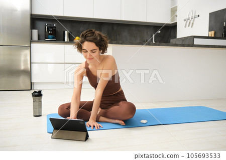 Portrait of smiling young woman, fitness instructor on rubber yoga mat, joins online training class from tablet, listens video in wireless headphones, workout at home on floor 105693533