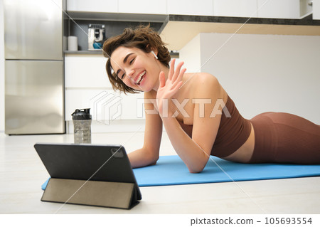 Sport and lifestyle. Young fitness woman, gym instructor waves hand at tablet, joins online video class, yoga and mindfulness workout, training from home indoors Sport and lifestyle. Young fitness woman, gym instructor waves hand at tablet, joins online video class, yoga and mindfulness workout, training from home indoors 105693554