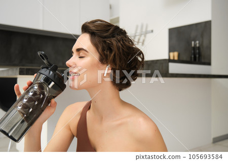 Smiling active young woman in sportswear, sits on yoga mat with water bottle, drinks stays hydrated during workout training session from home, fitness in the house on floor 105693584