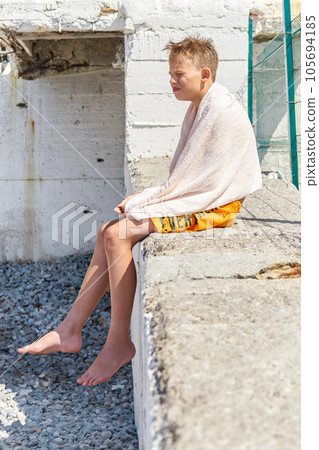 Preteen schoolboy in towel sitting on sunlit pier on beach 105694185