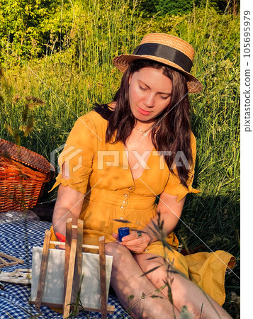 woman in a sunny yellow dress and hat paints at a picnic. 105695979