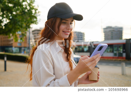 Red haired girl in casual clothes and black cap standing with mobile phone in the street. 105698966