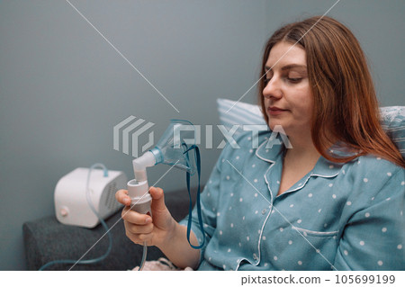 Young sick woman putting on nebulizer mask on face to make inhalation, using an inhaler at home. Medical inhaler emergency equipment, asthmatic attack 105699199