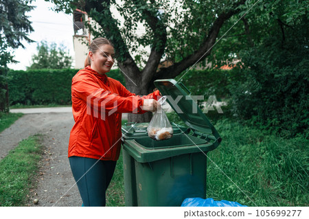 Caucasian woman sorting garbage, throwing a used bag with trash in a small recycle bin at outdoor near a home Caucasian woman sorting garbage, throwing a used bag with trash in a small recycle bin at outdoor near a home 105699277