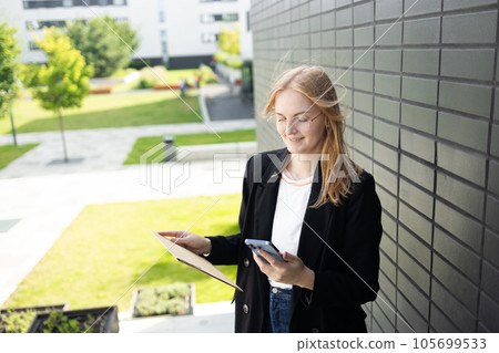 Portrait of corporate woman answers phone call while walking to the office building. Businesswoman calling someone, holding documents. 105699533