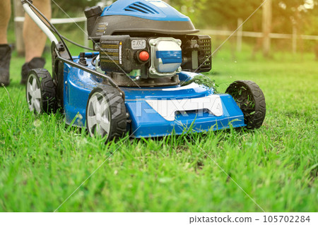 Lawn mower cutting grass. Small grass cuttings fly out of lawnmower. Grass clippings get spewed out of a mower pushed around by landscaper. CloseUp. Gardener working with mower machine. Mowing lawns Lawn mower cutting grass. Small grass cuttings fly out of lawnmower. Grass clippings get spewed out of a mower pushed around by landscaper. CloseUp. Gardener working with mower machine. Mowing lawns 105702284