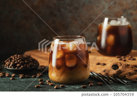 Ice coffee in a tall glass with cream poured over, ice cubes and beans on a dark concrete table. 105704359
