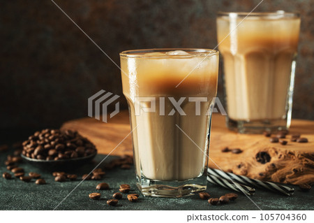 Ice coffee in a tall glass with cream poured over, ice cubes and beans on a dark concrete table. Ice coffee in a tall glass with cream poured over, ice cubes and beans on a dark concrete table. 105704360