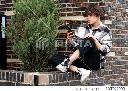 young man reads something on the phone while sitting on the brick curb 105704903