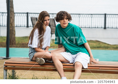 two young people, a guy and a girl, are sitting on a bench in a coastal park 105704943