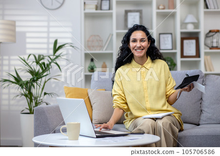 Portrait of beautiful woman at home, businesswoman smiling and looking at camera sitting on sofa in living room at home, woman paying bills using laptop, homework paper work with budget. Portrait of beautiful woman at home, businesswoman smiling and looking at camera sitting on sofa in living room at home, woman paying bills using laptop, homework paper work with budget. 105706586