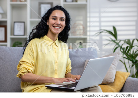 Portrait of young beautiful hispanic woman at home with laptop, woman smiling and looking at camera sitting on sofa in living room at home. Portrait of young beautiful hispanic woman at home with laptop, woman smiling and looking at camera sitting on sofa in living room at home. 105706992