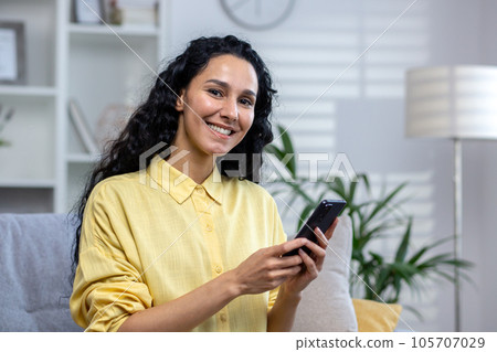 Portrait of a young beautiful hispanic woman at home, smiling and looking at the camera, holding a phone in her hands, using an application on a smartphone, browsing the Internet, typing a message. Portrait of a young beautiful hispanic woman at home, smiling and looking at the camera, holding a phone in her hands, using an application on a smartphone, browsing the Internet, typing a message. 105707029