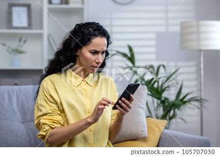 Upset and disappointed serious thinking woman alone at home using app on phone close up, hispanic sitting on sofa in living room at home. Upset and disappointed serious thinking woman alone at home using app on phone close up, hispanic sitting on sofa in living room at home. 105707030