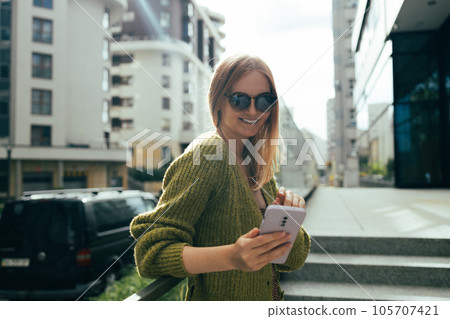 Portrait smiling young Caucasian beautiful woman wearing sunglasses sitting outside in city looking at mobile phone 105707421