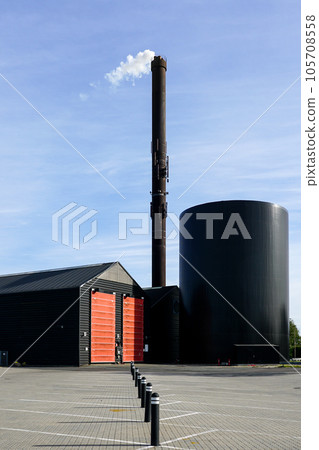 Modern factory with a tall rusty chimney from which comes out white steam on blue sky background 105708558