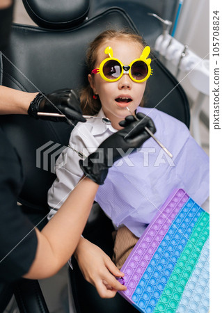 Vertical cropped shot of unrecognizable pediatric dentist examining teeth of adorable little child girl in funny eyeglasses lying on dental chair in dentistry clinic, playing with colorful pop it toy Vertical cropped shot of unrecognizable pediatric dentist examining teeth of adorable little child girl in funny eyeglasses lying on dental chair in dentistry clinic, playing with colorful pop it toy 105708824