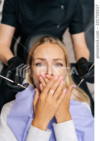 Vertical portrait of afraid blonde female patient closing mouth with hands, preventing examining of teeth, because of fear, looking at camera with scared expression. Healthcare, dental health concept. Vertical portrait of afraid blonde female patient closing mouth with hands, preventing examining of teeth, because of fear, looking at camera with scared expression. Healthcare, dental health concept. 105708837