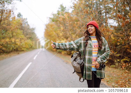Young female hitchhiker by roadside among autumn forest during fall season. Travel woman hitchhiking 105709618