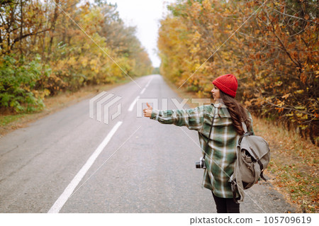 Young female hitchhiker by roadside among autumn forest during fall season. Travel woman hitchhiking Young female hitchhiker by roadside among autumn forest during fall season. Travel woman hitchhiking 105709619