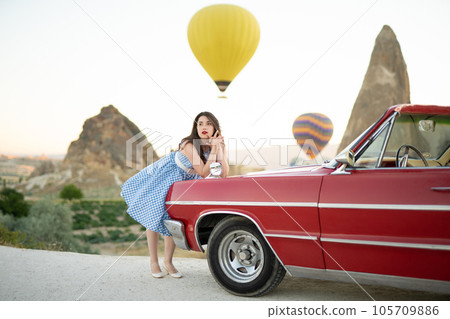 beautiful girl in retro style posing near a vintage red cabriolet car on background of balloons in Cappadocia. beautiful girl in retro style posing near a vintage red cabriolet car on background of balloons in Cappadocia. 105709886