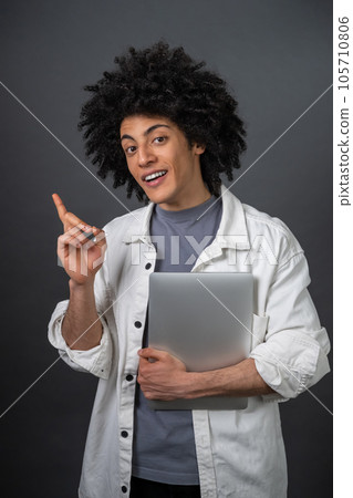 Curly-haired young guy with laptop smiling happily and pointing with his finger 105710806