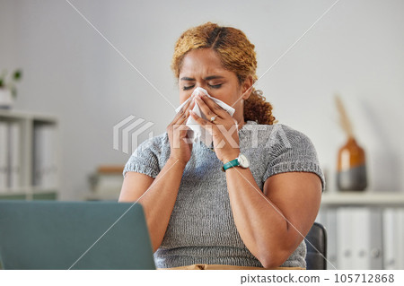Sick, ill, or unwell business woman suffering with a cold, flu or sinus infection and sneezing or blowing her nose with a tissue. Young female remote working on her laptop from the home office 105712868