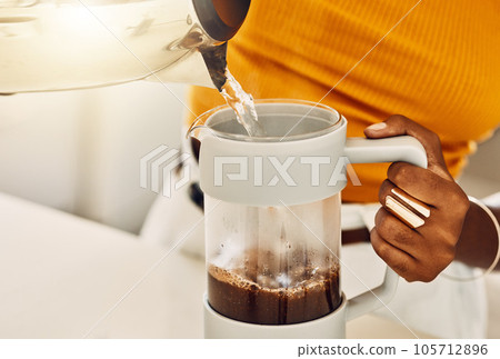 Female hands making morning coffee to enjoy with breakfast, pouring hot water from kettle in a kitchen. Closeup of casual, black woman preparing or brewing tea or espresso beverage at home. Female hands making morning coffee to enjoy with breakfast, pouring hot water from kettle in a kitchen. Closeup of casual, black woman preparing or brewing tea or espresso beverage at home. 105712896