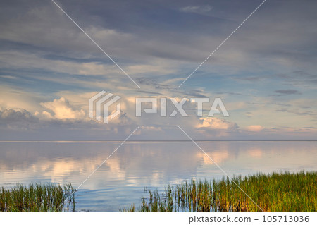 A beautiful calm and quiet view of lake water with a cloudy sky reflection and green grass on the bank. The landscape of a magical horizon with copy space outdoors in nature or a peaceful environment A beautiful calm and quiet view of lake water with a cloudy sky reflection and green grass on the bank. The landscape of a magical horizon with copy space outdoors in nature or a peaceful environment 105713036