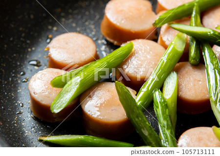 A cooking scene where frankfurter sausage and garlic sprouts are fried. 105713111