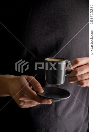 A man's hand holds a cup of hot espresso coffee against the background of a black T-shirt. A man's hand holds a cup of hot espresso coffee against the background of a black T-shirt. 105715283