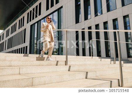 Stylish woman entrepreneur talking phone while crossing on modern building background with coffee Stylish woman entrepreneur talking phone while crossing on modern building background with coffee 105715548