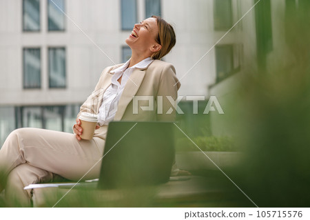 Smiling business woman sitting on bench and drinking coffee during break time near office building 105715576