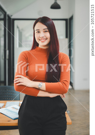 Portrait of young smiling woman looking at camera with crossed arms. Happy girl standing in creative office. Successful businesswoman standing in office with copy space. 105721515