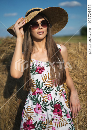 beautiful glamorous girl in a dress and a straw hat on a field near a haystack. 105724423