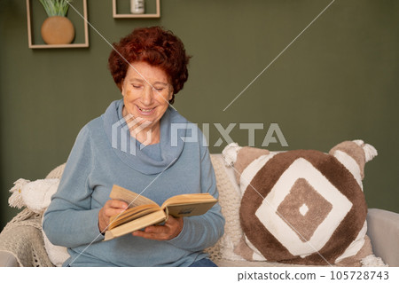 Portrait of a happy modern senior woman in a blue sweater with red hair who sits on the sofa during the day in the living room, smiles and reads a book Portrait of a happy modern senior woman in a blue sweater with red hair who sits on the sofa during the day in the living room, smiles and reads a book 105728743
