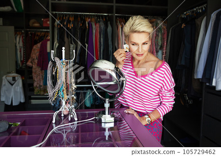 Mature and beautiful woman with short, blonde hair trying on accessories, earrings, while looking into the mirror. Model aiming for style suitable for women of middle age 105729462