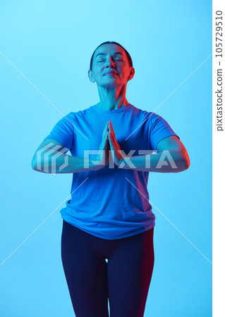 Beautiful mature woman standing in yoga pose, relaxing body and mind against blue studio background in neon light Beautiful mature woman standing in yoga pose, relaxing body and mind against blue studio background in neon light 105729510
