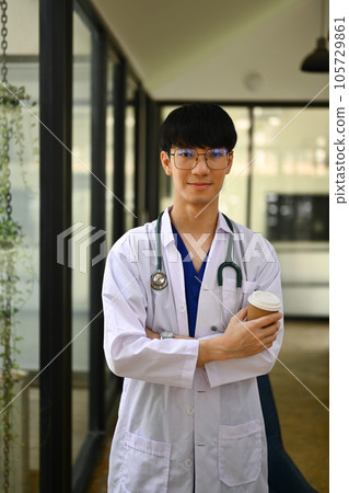 Portrait of male general practitioner in white coat standing near window and looking at camera Portrait of male general practitioner in white coat standing near window and looking at camera 105729861