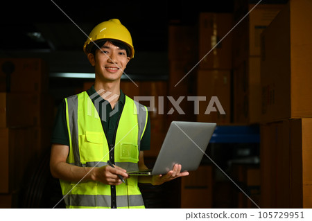 Smiling male manager wearing hardhat and reflective jacket standing between shelves with cardboard boxes 105729951