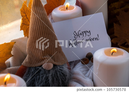 HAPPY HOLLOWEEN text greeting card concept Celebrating halloween autumn holidays at cozy home on the windowsill Hygge aesthetic atmosphere Autumn leaves gnome spices and candle on knitted white 105730578