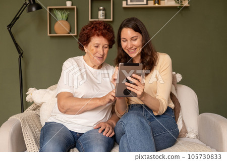 Portrait of a modern happy grandmother and granddaughter who sit in the living room during the day and look at the tablet. Woman with a smile and interest touches a gadget with her hand 105730833